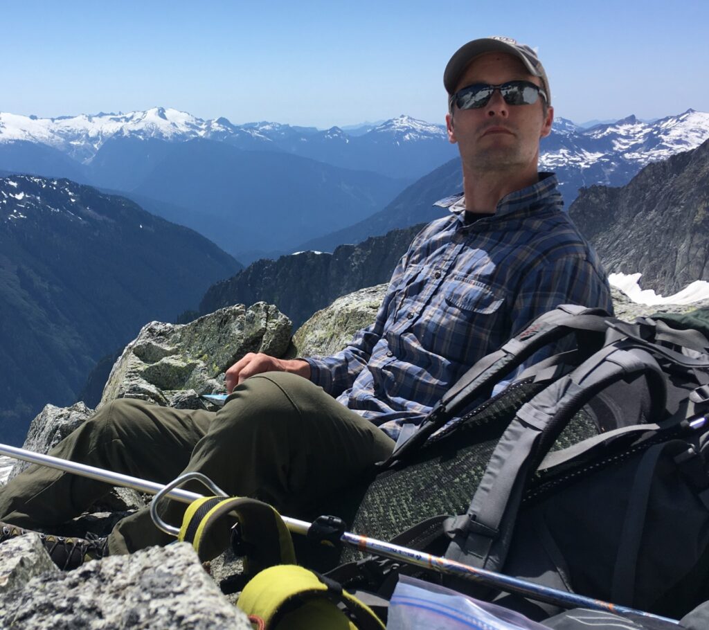 Dan sits on top of the mountain with his backpack and hiking poles