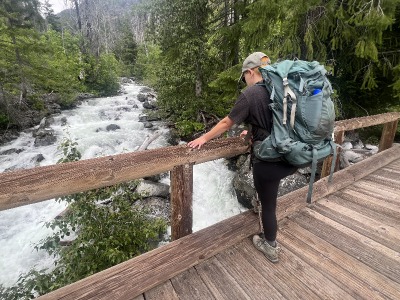 A hiker carrying a large blue backpack and standing on a wooden bridge looking at a river below.