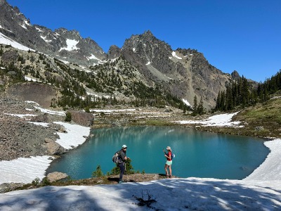 Two hikers standing next to a turqouise colored lake with mountains and blue skies.