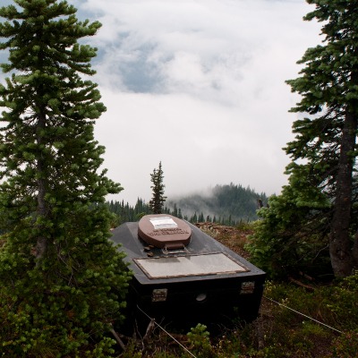 a large black box with a toilet seat between two trees with a foggy mountain view.