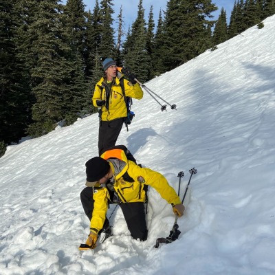 Two men in yellow jackets standing on a snowy slope with hiking poles and avalanche safety gear.