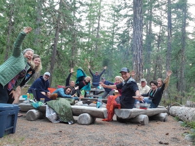 A group photo of 13 people sitting on wooden benches by the campfire with their hands up.