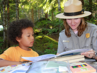 A boy sitting with a park ranger while reading a book.