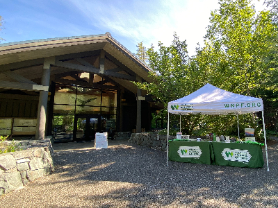 Photo of a white canopy with a green table underneath, set up next to a large wooden building.