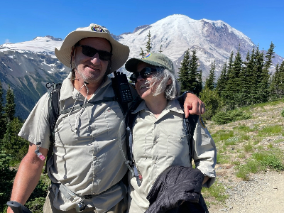 A man and woman dressed in khaki hiking clothes smiling in front of Mount Rainier