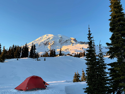 A red tent pitched in a snow covered meadow with a large mountain in the background with the sun shining on it.