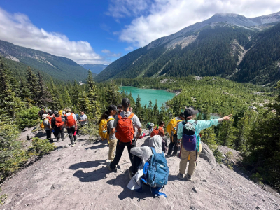 a large group of hikers standing on a rock, overlooking a turquoise lake.