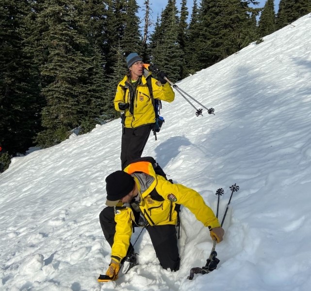 Two men in yellow jackets holding avalanche tranceivers and standing in a snowy hill.