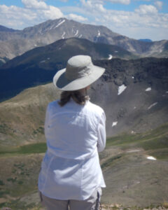 A woman facing away from the camera looking at mountains. She is wearing a white shirt and gray hat.