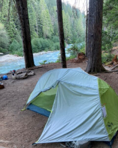 Photo of a green and white tent pitched in a campsite by a blue river.