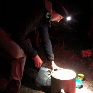 A man in the dark wearing a headlamp with camp kitchen gear on the ground.