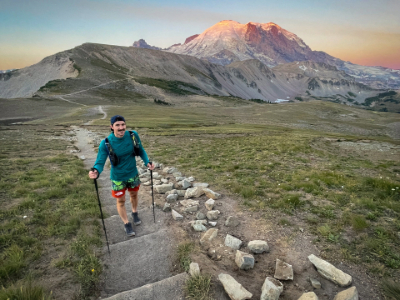 A man standing on a trail holding hiking poles at sunrise with a mountain behind him.