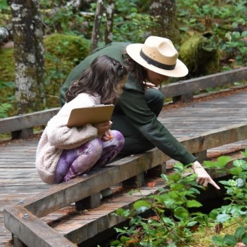 A park ranger and a young student on a boardwalk pointing at a plant.