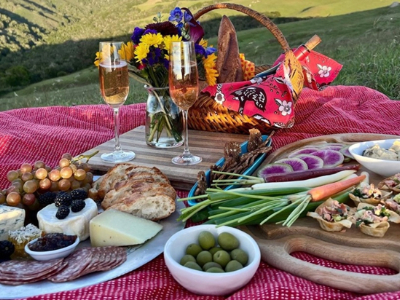 A plate of meat, cheese, crackers, and champagne spread out on a red blanket.