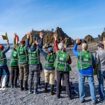 8 volunteers standing on a beach wearing green vests with their hands up