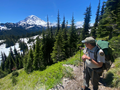 A hiker standing on a trail looking to his right at a blue sky and Mount Rainier in the distance.