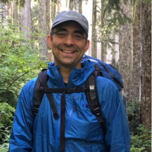 A man wearing a blue raincoat, backpack, and hat, standing in a forest.