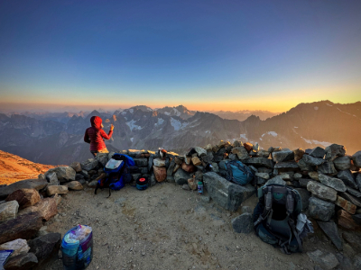 A person wearing a red jacket taking a photo of sunset while sitting on a stack of rocks.