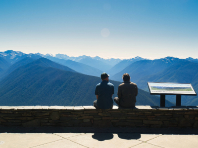 Two people sitting on a ledge, looking at blue skies and expansive views of Olympic mountains. 