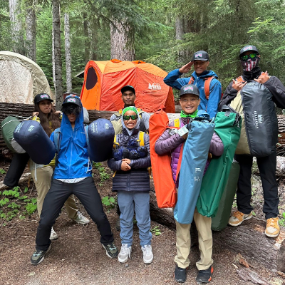 Seven teenagers holding sleeping bags, tents, and camp chairs while posing infront of an orange tent in the woods.